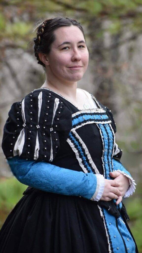 Sophia Berkeley, femme presenting, wearing a Tudor style black wool gown with blue and silver accents.